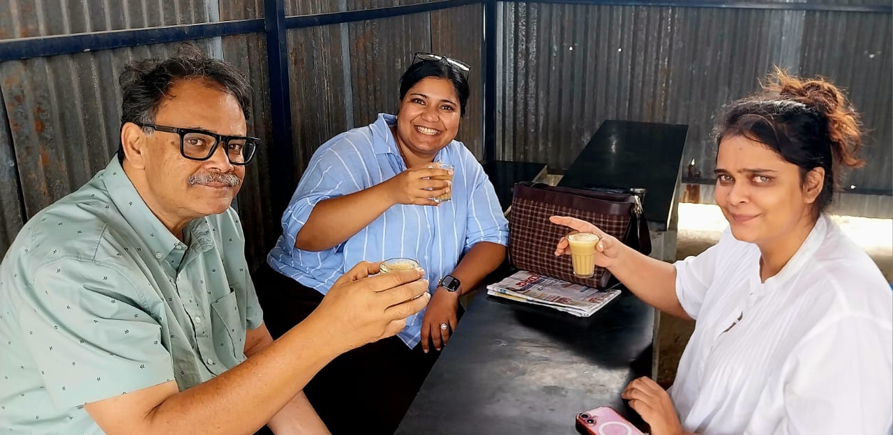 Three people raise small glasses of tea across a table in a corrugated-sheet tea shed